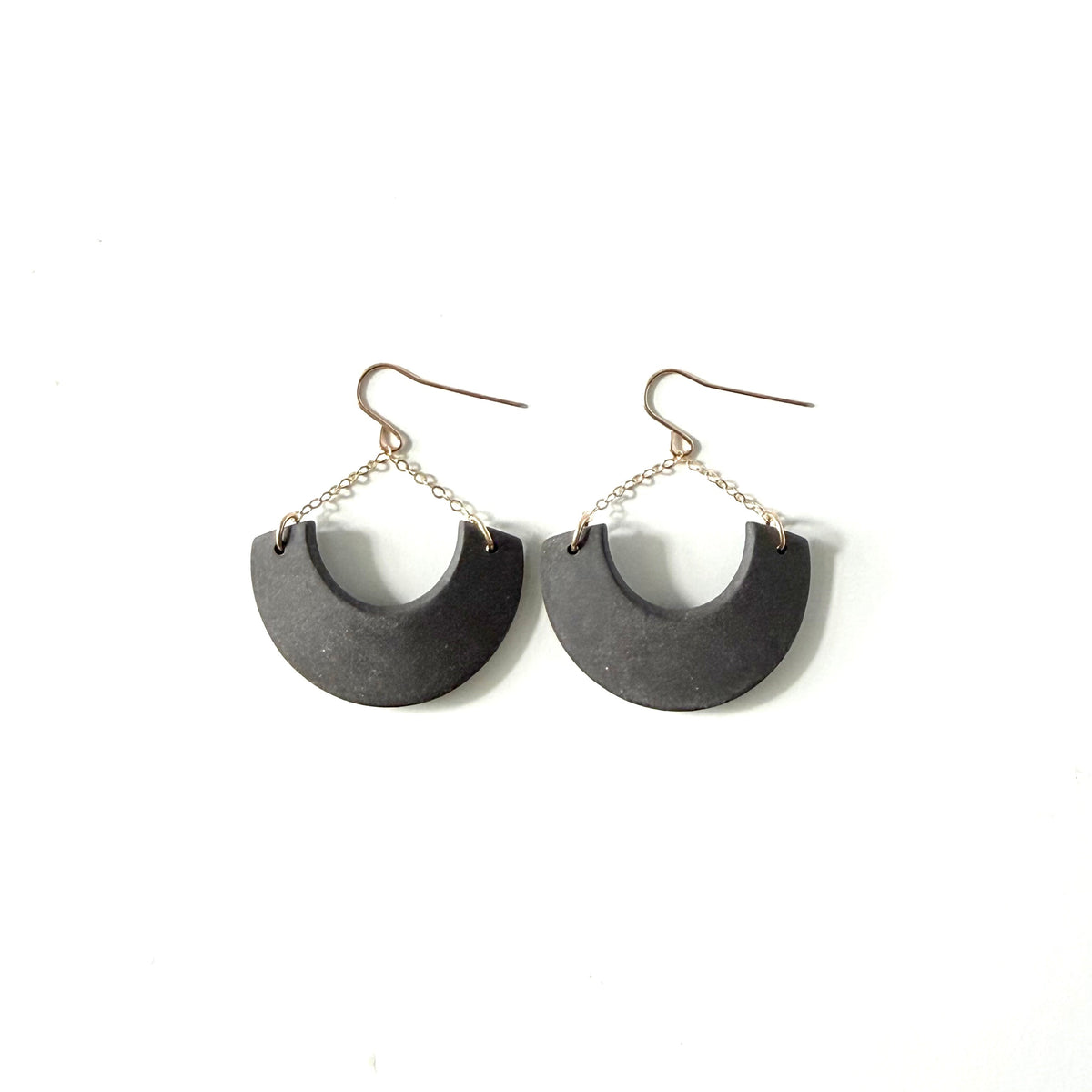 A pair of handmade semi-circular drop earrings with dark charcoal black textured discs suspended from thin gold chains and fishhook wires, shown on a plain white background.