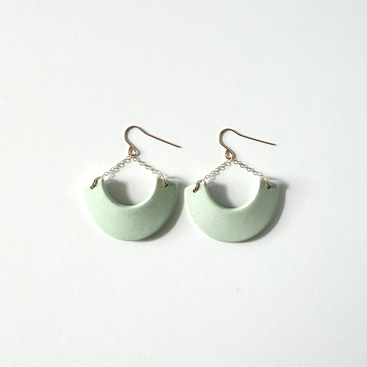 A pair of light mint green crescent-shaped drop earrings with gold-tone fishhook wires and short chain links, displayed on a clean white background.